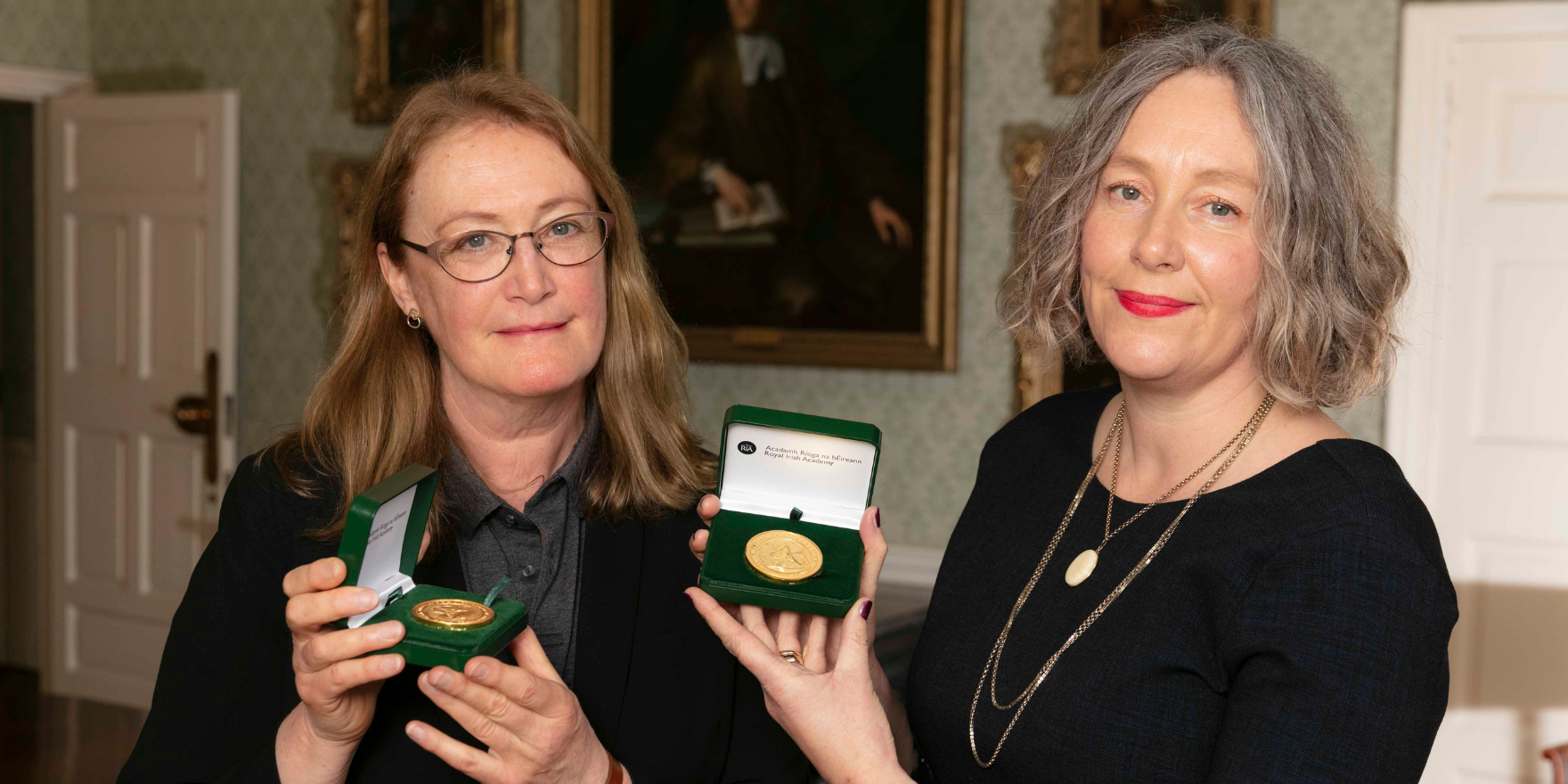 Two women (Ruth M.J. Byrne MRIA on the left and Jennifer C. McElwain MRIA on the right) pose for a photograph (from the shoulders up) with their RIA Gold Medals in a green case, held in both their hands. Ruth M.J. Byrne MRIA is wearing glasses and has shoulder-length strawberry blonde hair. She is wearing a grey shirt and black blazer. Jennifer C. McElwain MRIA has silver curly hair and wears red lipstick. She is wearing black and wears gold jewellery. A painting hangs on a wallpapered wall in the background of the lavish room they stand in.
