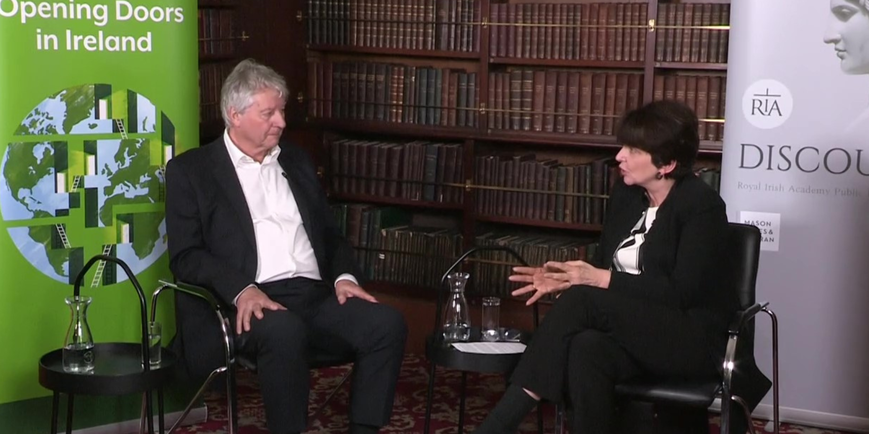 A man with grey short hair (left) and a woman with dark short hair (right) sit facing each other in office chairs, they are in conversation. Both are wearing suits. A bookcase lined with leather bound books is behind them, two pull up banners stand on either side of the chairs.