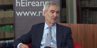 A man with grey hair wearing a black suit and blue shirt and tie is seated and is in mid conversation with someone to the right of the image