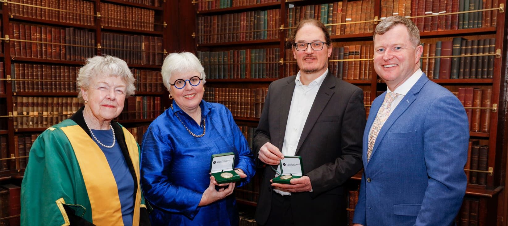Four people standing in a row in front of a walled book shelf smiling at the camera. A woman [Dr Mary Canning, President of the Royal Irish Academy (2020-2023)] stands to the left, a man (Jonathan N. Coleman MRIA, 2023 RIA Gold Medallist in the Physical and Mathematical Sciences) and a woman (Jane Ohlmeyer MRIA, 2023 RIA Gold Medallist in the Humanities) stand in the centre, both of them are holding a green case containing a gold medal. A man (Senator Malcolm Byrne, Fianna Fáil's Spokesperson on Further and Higher Education, Research, Innovation and Science) stands on the right after presenting the Gold Medals to the winners.