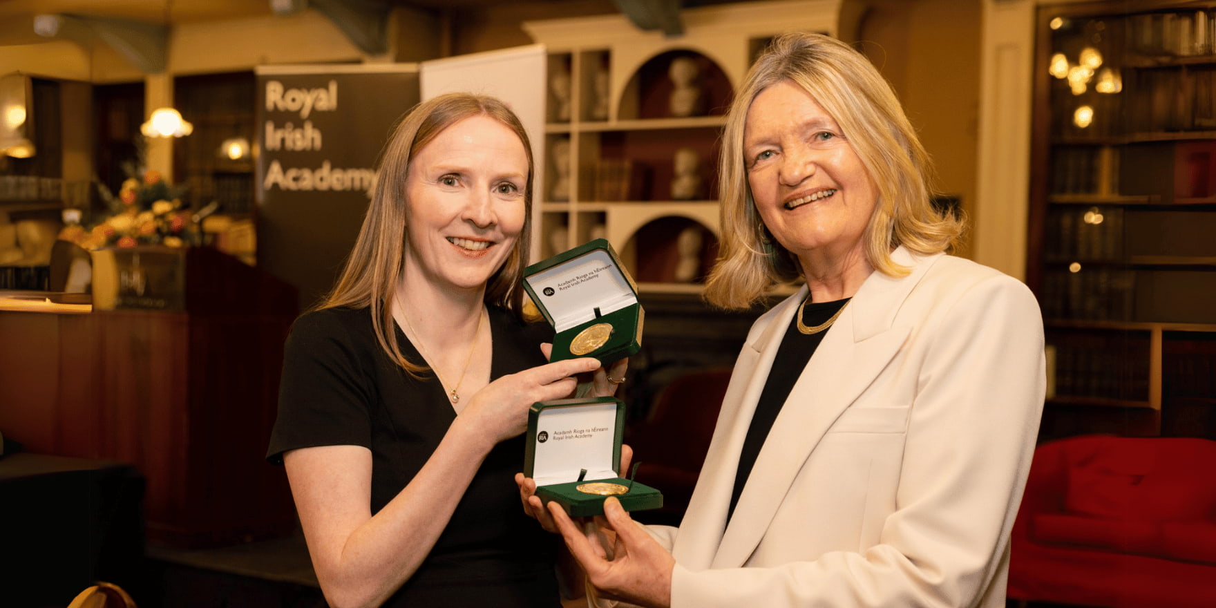 Two females (Máire O’Neill MRIA and Brigid Laffan MRIA) one with auburn hair, the other with blonde smiling at the camera holding two RIA Gold Medals showcased in a green box.
