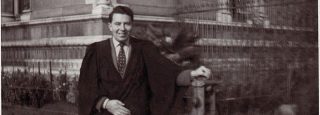 Black and white photograph of R.J. (Bob) Hunter outside Trinity College Dublin c. 1960. He has dark hair and is smiling, dressed in a robe with one hand resting on a railing.