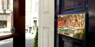 A close-up view of a dark wooden door with a polished brass plaque that reads 'Royal Irish Academy.' The door is slightly open, and through the doorway, a city street with blurred buildings is visible in the background