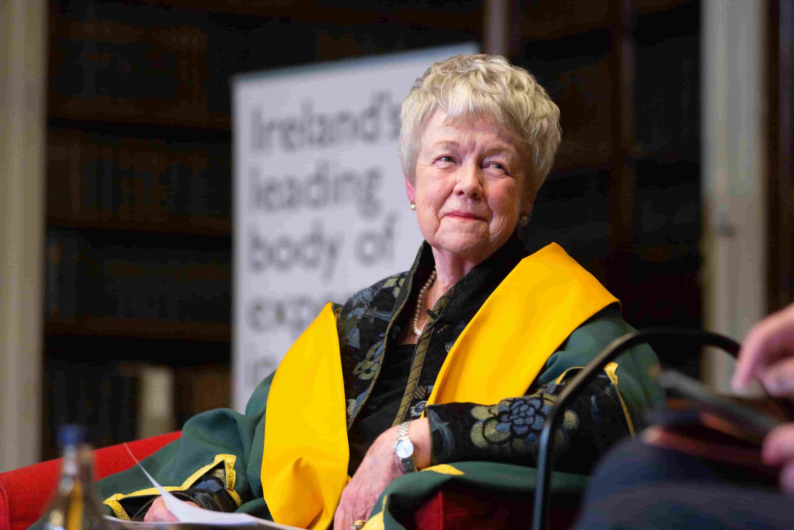 Image of a woman with white cropped hair sitting in a chair. She is wearing yellow and green ceremonial robes and smiling toward someone to the right.