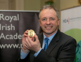 A man (Alan Smeaton MRIA) smiles and poses for a photograph with his RIA Gold Medal cupped in both hands. He has grey hair and is wearing a blue shirt, blue tie and dark coloured suit jacket. He is photographed from the arms up. There are large banners in the background of the photograph one reads HEA and the other reads Royal Irish Academy in large letters.