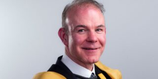 A headshot of a man with grey hair, against a grey background, wearing the yellow and green robes of the Royal Irish Academu
