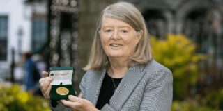 A woman (Mary E. Daly) with shoulder length grey hair poses for a photo with her RIA Gold Medal.