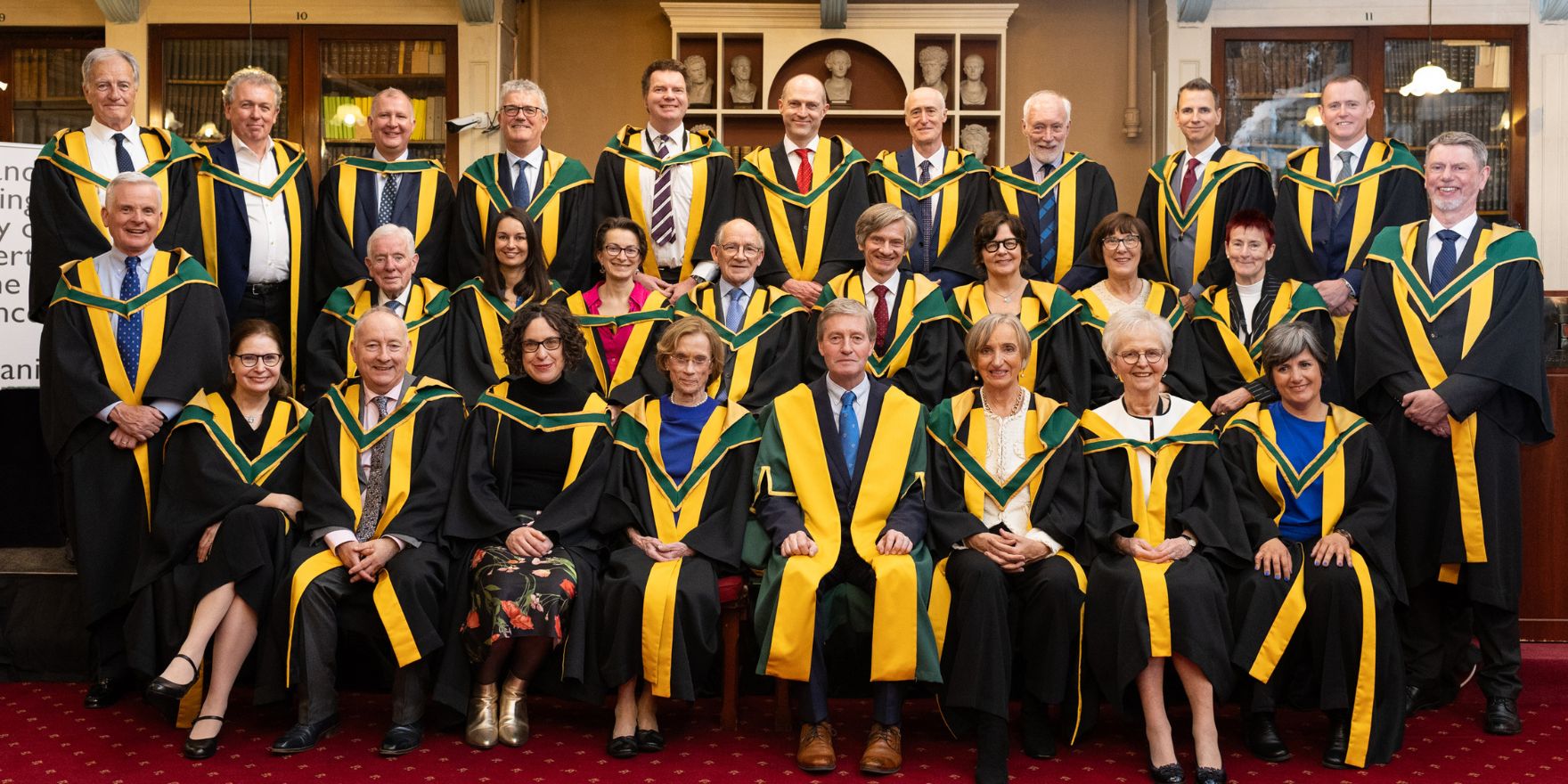 A group of 27 women and men, standing at the back row, with the two rows in front seated, on a stage, all dressed in yellow and green robes, the official robes of the Royal Irish Academy.