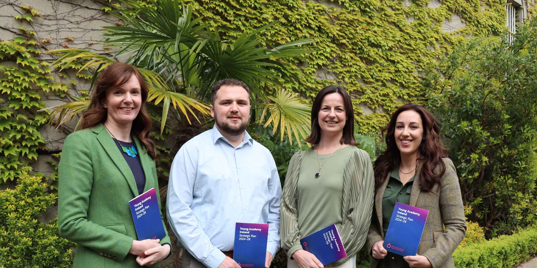 Four members of Young Academy Ireland (YAI), three woman and one man, holding printed copies of the YAI strategic plan, standing in front of a wall, covered in greenery