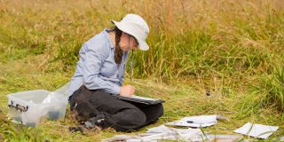 A woman wearing a hat, sits in a grassy field making notes in a notebook