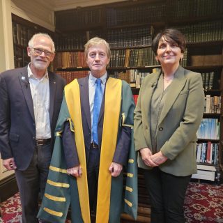 Three individuals stand before a bookcase, with one man proudly wearing a presidential robe