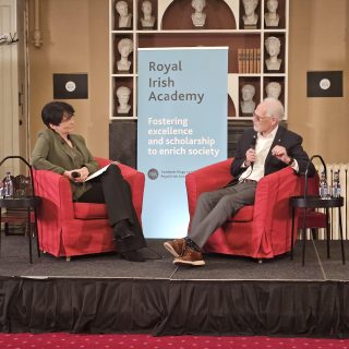 Two individuals seated on stage at the Royal Institute of Irish Academy, engaged in discussion.