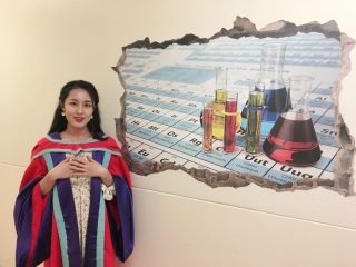 Woman in graduation robes standing in front of a mural featuring chemistry lab equipment.